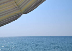 Blick auf das Mittelmeer. Der Himmel ist blau, in der linken oberen ecke sieht man angeschnitten einen Sonnenschirm mit Streifenmuster.