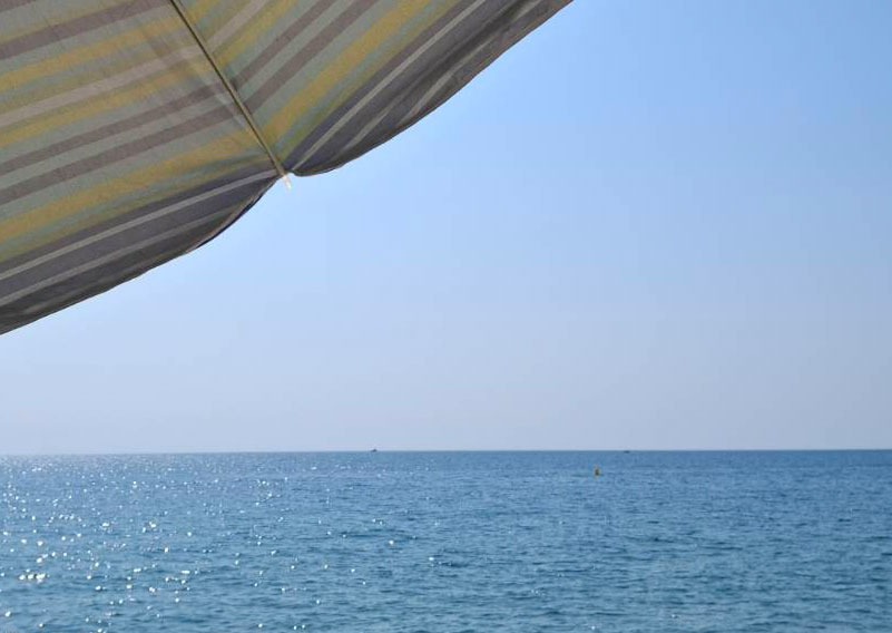 Blick auf das Mittelmeer. Der Himmel ist blau, in der linken oberen ecke sieht man angeschnitten einen Sonnenschirm mit Streifenmuster.